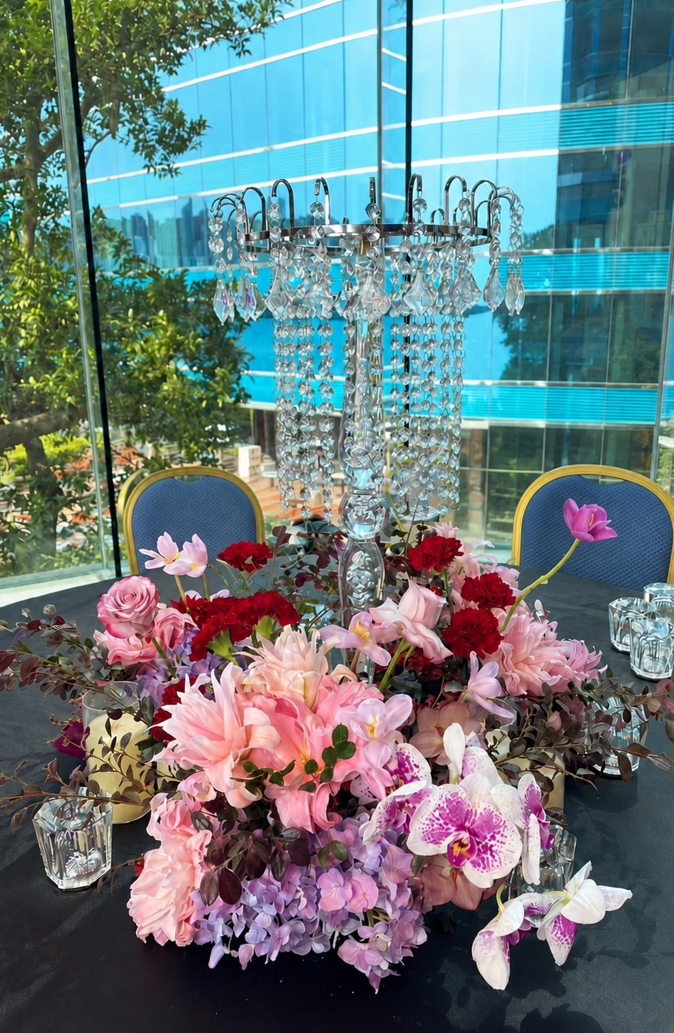 水晶吊飾燭台下的粉色百合與蝴蝶蘭桌花，後方為玻璃帷幕與藍色建築