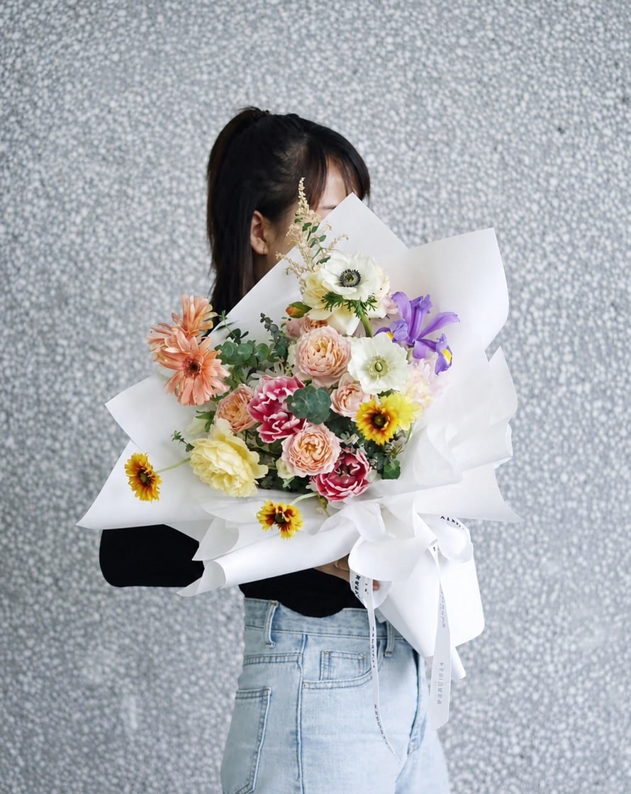 橘色非洲菊、小向日葵與紫色鳶尾等混合花束,白色包裝紙,灰色水泥牆背景